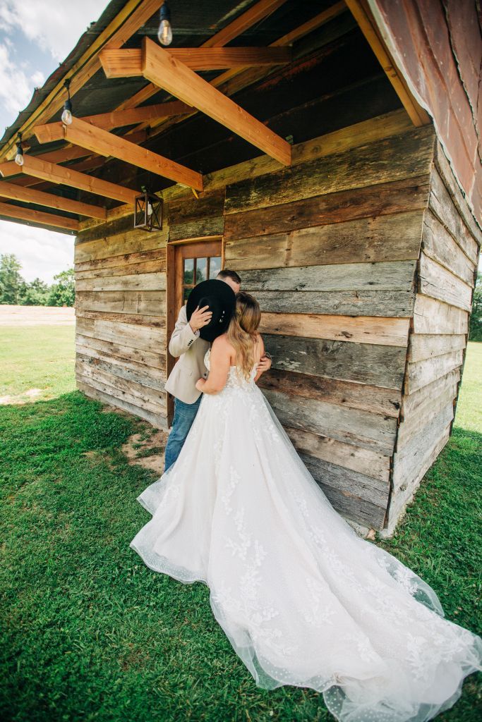 bride and groom kiss behind his hat next to a rustic wooden cabin on their wedding day