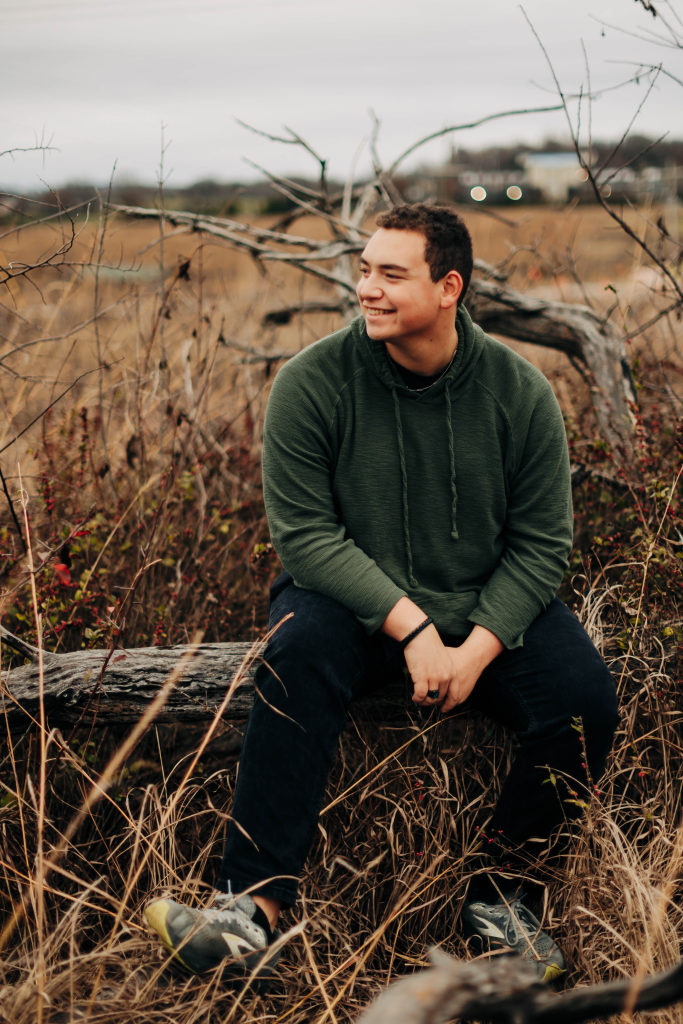 A Harrison senior sits on a tree log with dry grass around him and smiles as he looks at the horizon