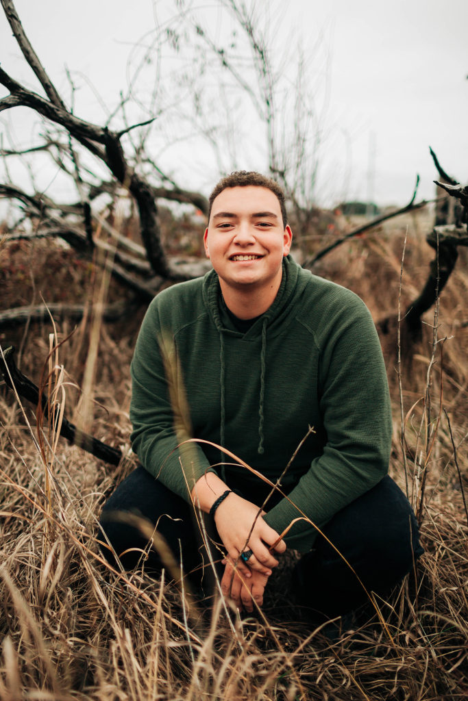 A senior sits amidst dry grass and smiles at the camera