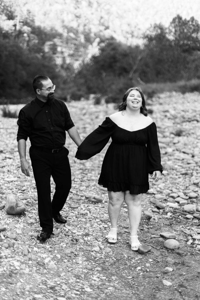 a couple walk together and laugh on a rocky riverside during their engagement photos