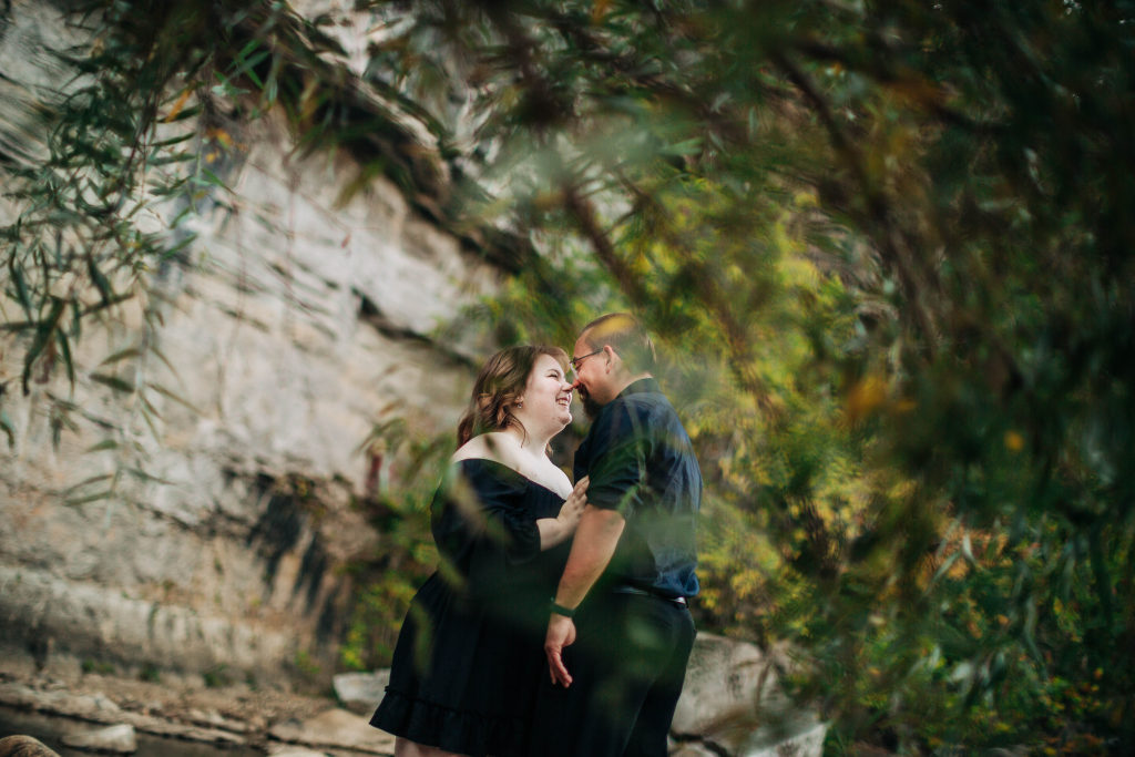 a couple show their joy as they look into each others eyes in the background of a green tree and against a rocky wall