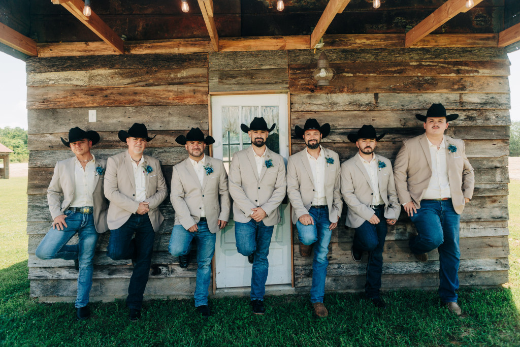 groomsmen pose with their stetson hats next to a rustic cabin in the ozarks