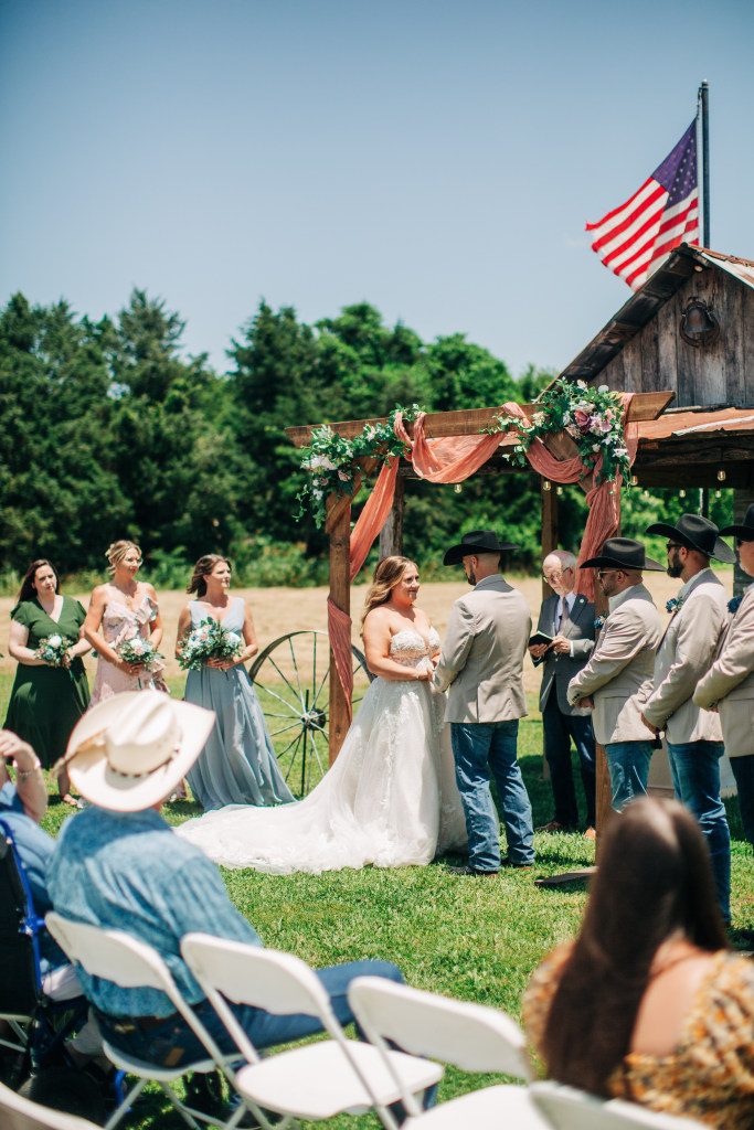 couple say their vows in the full sun at the White Barn with the bridal party surrounding them
