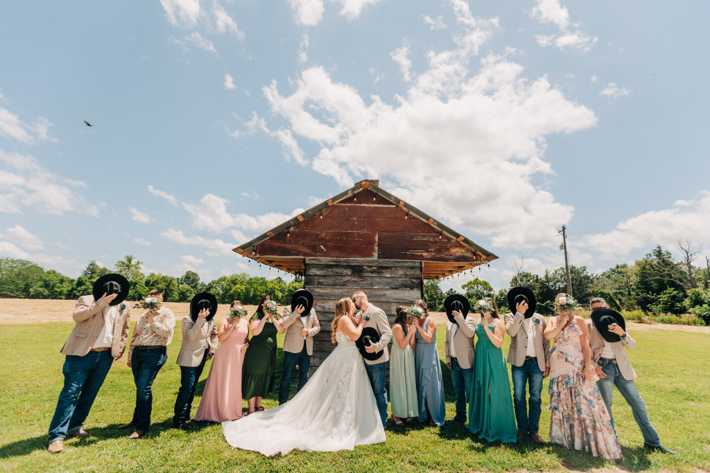bridal party hides behind bouquets and stetson hats while the bride and groom kiss