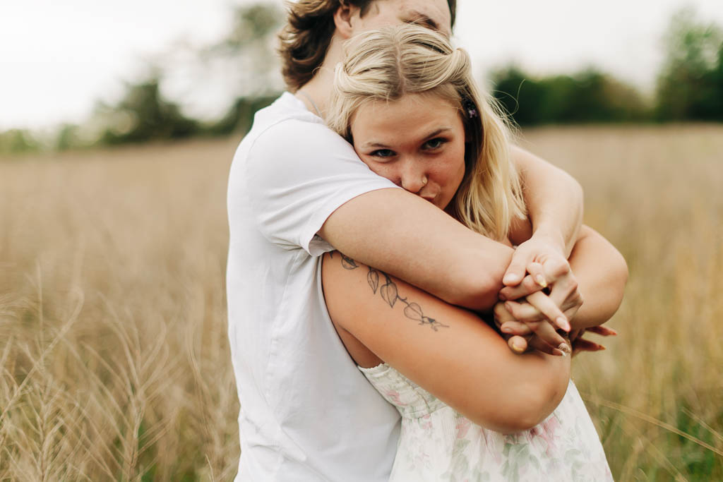 girl playfully kisses her boyfriend's arm while in his embrace in a grassy field hear Harrison AR