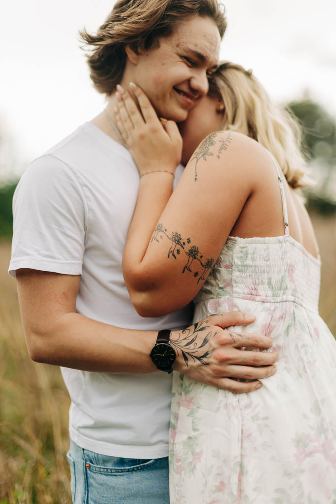 a guy and girl with cute tattoos embrace and smile in a grassy field
