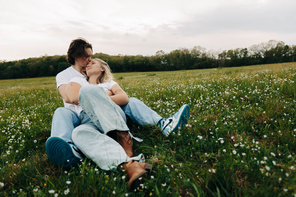 a girl relaxes into her boyfriend's embrace while seated in a country flower field
